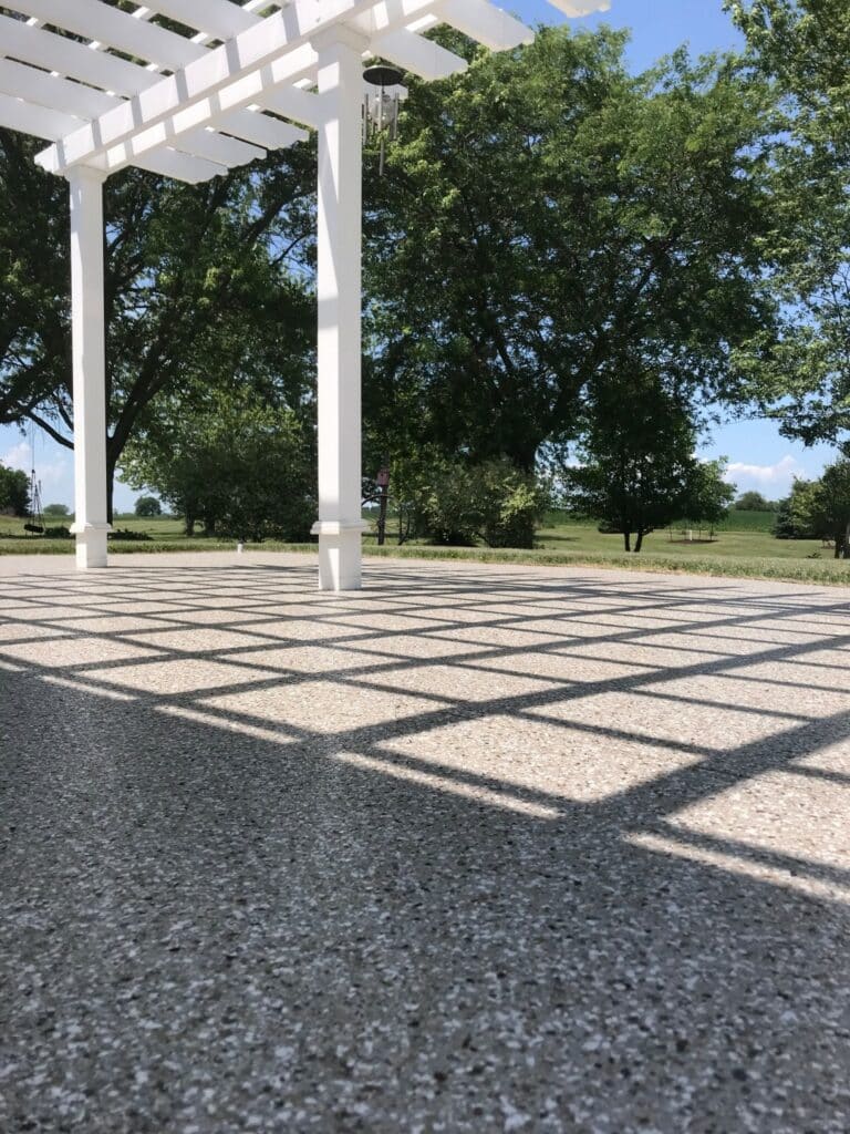 A white pergola casts shadows on the ground; surrounded by green trees under a clear blue sky, creating a peaceful outdoor setting.