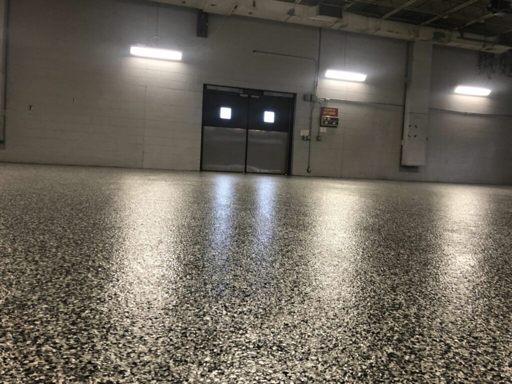 Empty industrial room with a speckled floor and overhead lighting. Walls are gray, featuring a metal double door and exposed ceiling pipes.