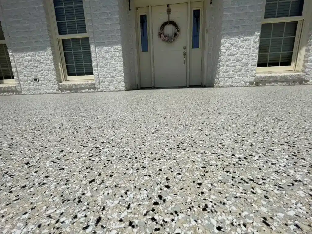 Textured concrete floor leading to a white brick building entrance with wreath-adorned door, flanked by windows with shutters.