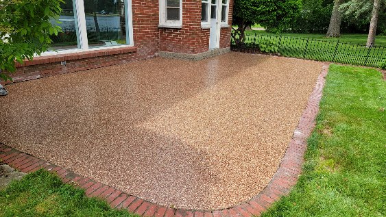 A freshly paved patio with a pebble-like surface adjoins a brick house, surrounded by green grass and trees, creating a serene backyard space.