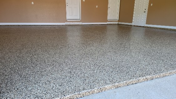 The image shows an empty garage with tan walls, a speckled epoxy floor, and white interior doors. No people or landmarks are present.