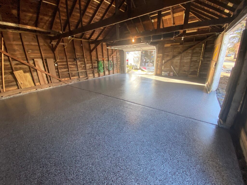 A spacious garage with exposed wooden beams and a newly finished epoxy floor, revealing open doors to a bright outside area.