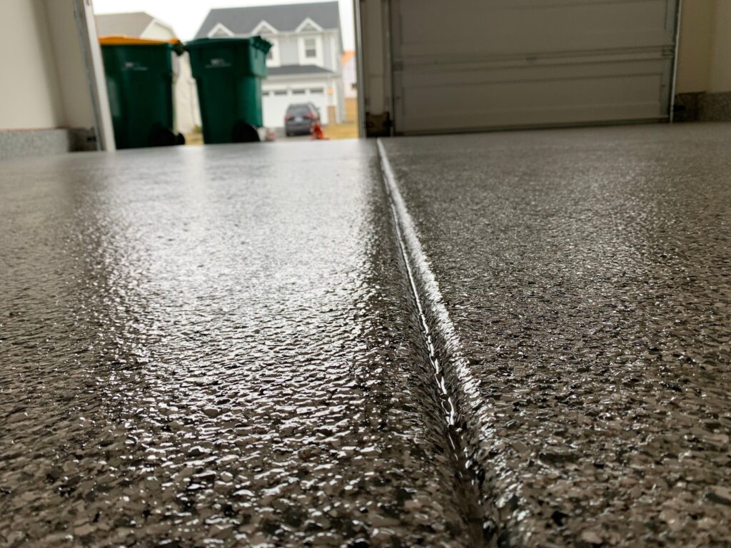 Close-up of a textured garage floor with a narrow groove, open garage door, two green bins, and a house in the background.