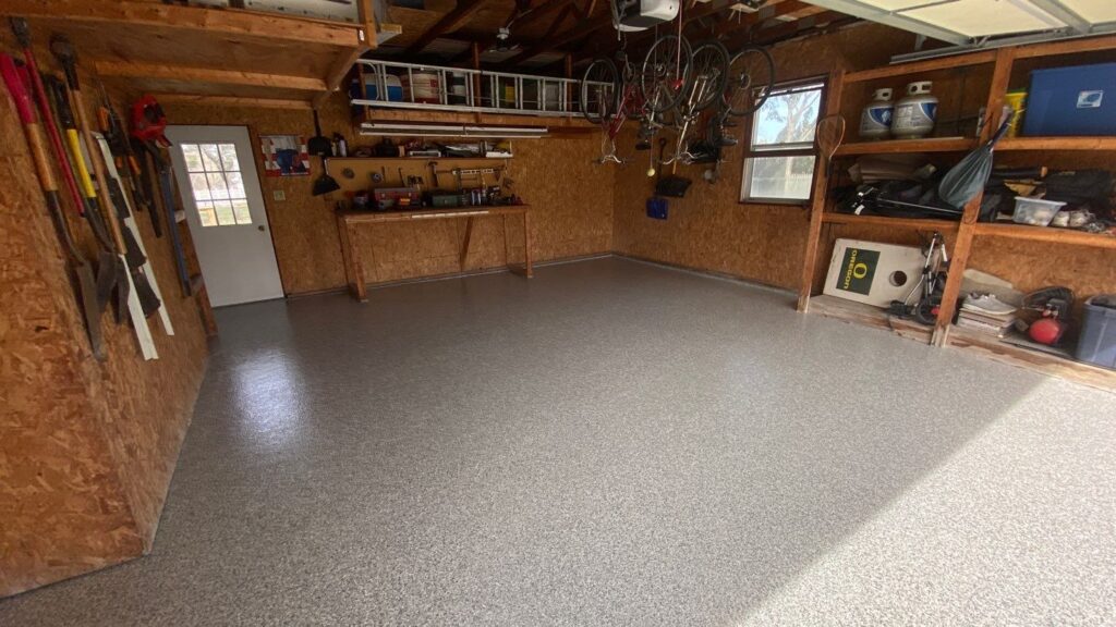 A tidy garage with a workbench, organized tools on shelves, hanging bicycles, and a solid grey floor, illuminated by natural light.