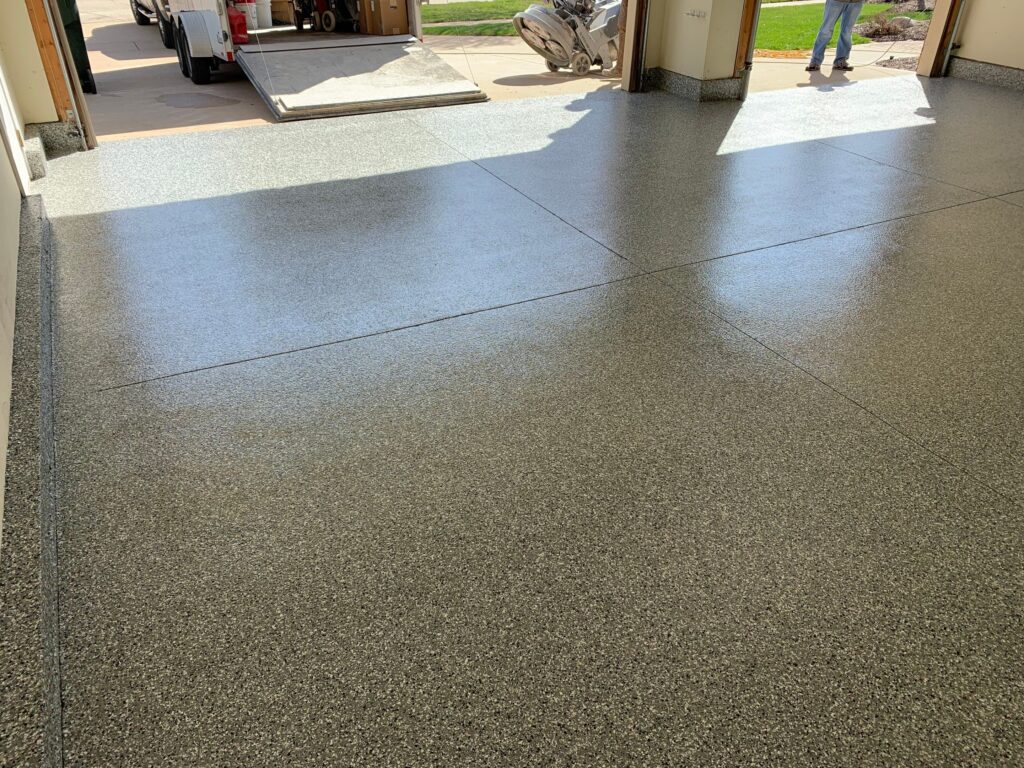 A newly finished epoxy garage floor gleams under sunlight. A person stands outside near a trailer in a tidy driveway.