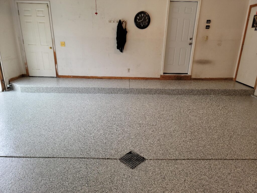 A clean garage with speckled floor coating, multiple white doors, a hanging black coat, and a wall clock in the background.