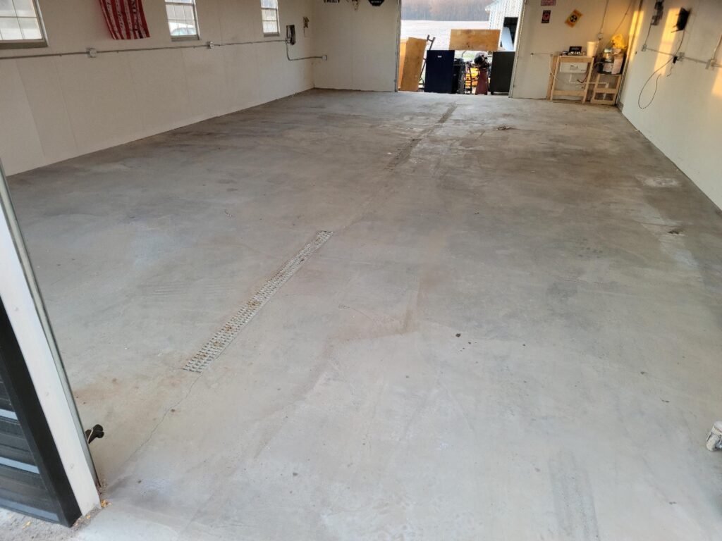 An empty garage with a concrete floor, white walls, wall-mounted shelves, and tools. Sunlight streams through open doors at one end.