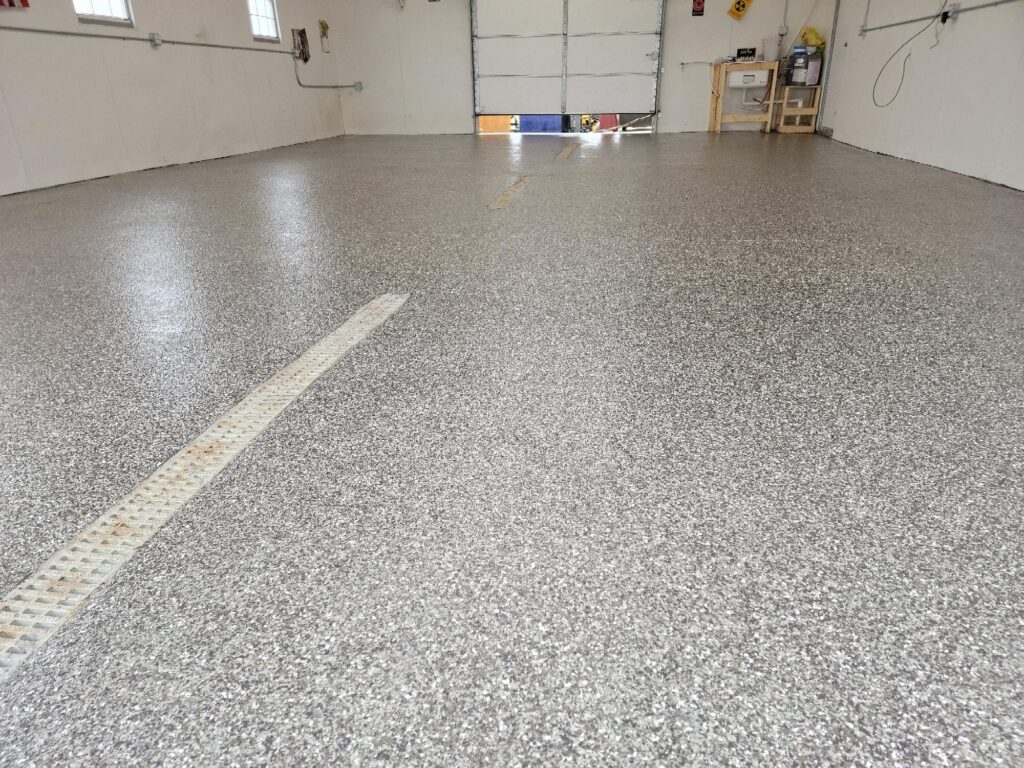 Clean garage with gray speckled epoxy flooring, bright lighting, garage door, workbench, and storage shelves. No people or cars present.