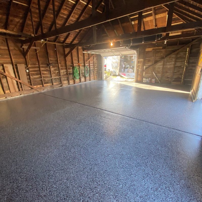 A spacious garage with exposed wooden beams and a newly finished epoxy floor, revealing open doors to a bright outside area.