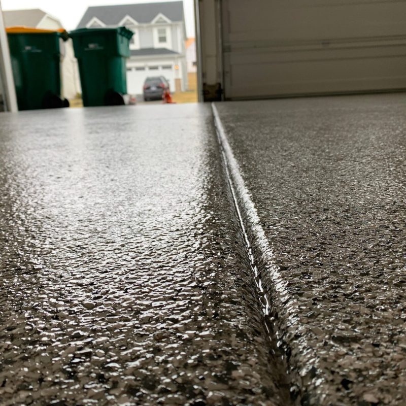 Close-up of a textured garage floor with a narrow groove, open garage door, two green bins, and a house in the background.