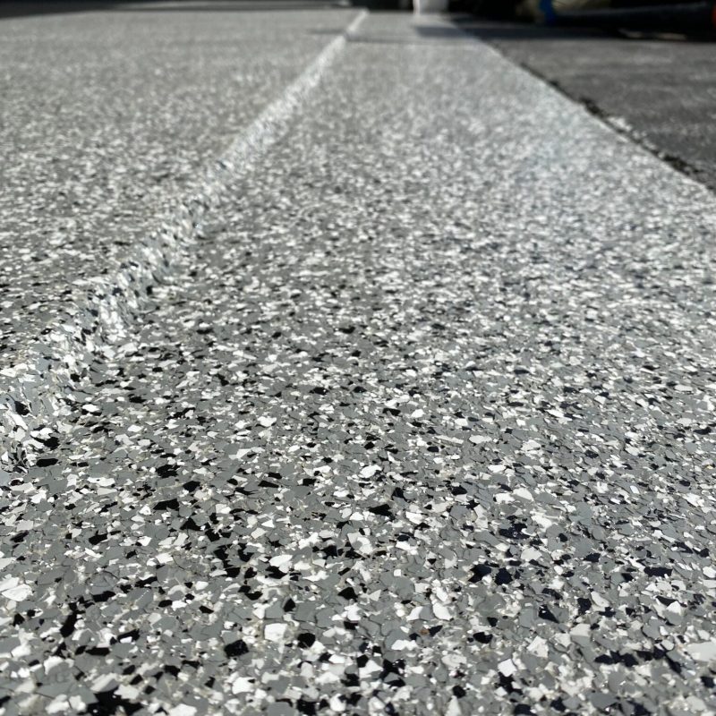 Close-up of a speckled, concrete garage floor. In the background, bags are stacked near an open garage door with partial sunlight.