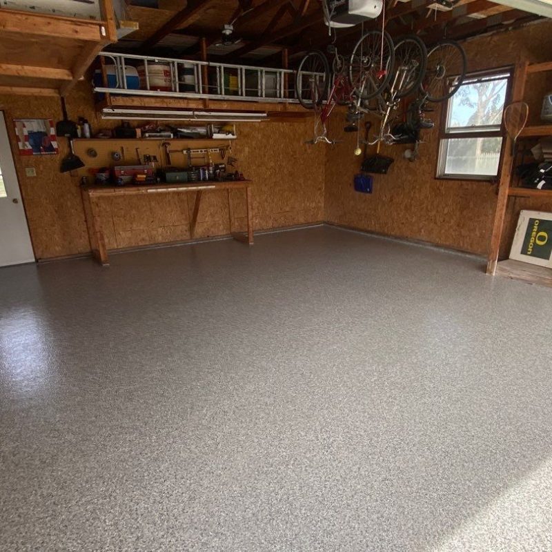 A tidy garage with a workbench, organized tools on shelves, hanging bicycles, and a solid grey floor, illuminated by natural light.