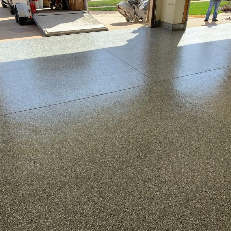 A newly finished epoxy garage floor gleams under sunlight. A person stands outside near a trailer in a tidy driveway.