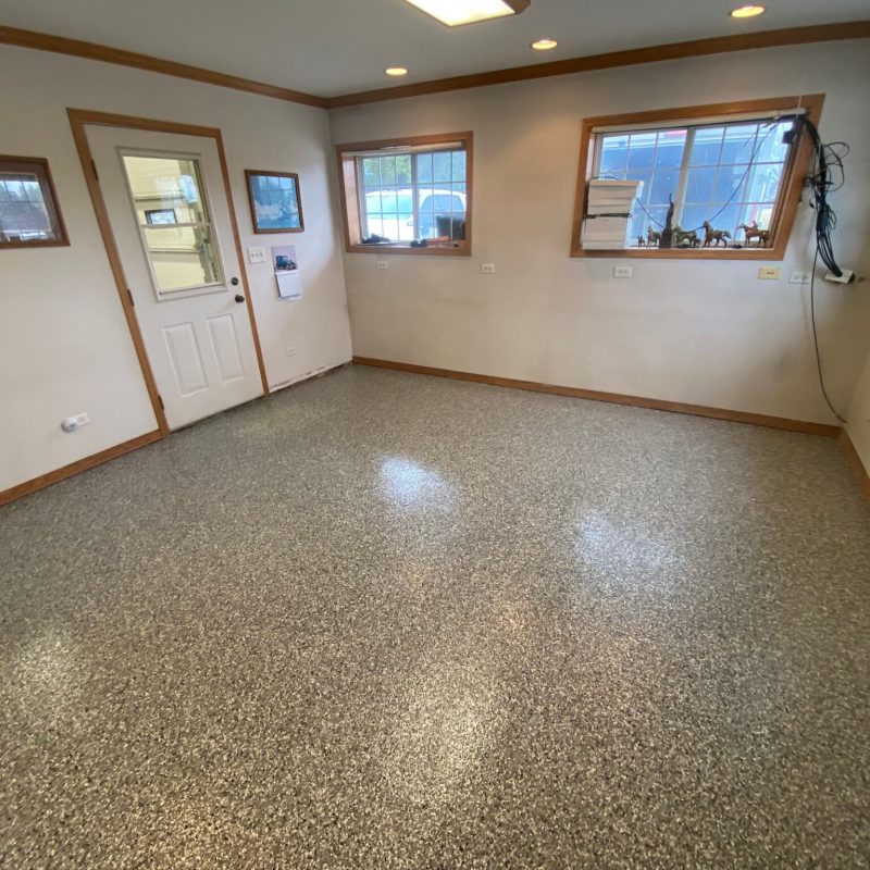 An empty room with speckled flooring, beige walls, framed pictures, and windows revealing outdoor views. Door and wall outlets are visible.